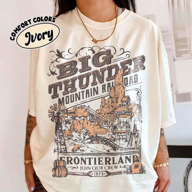 May include: An ivory short-sleeve t-shirt featuring a graphic print with the text BIG THUNDER MOUNTAIN RAILROAD FRONTIERLAND JOIN OUR CREW 1979. The illustration depicts a mountain landscape, a windmill, and barrels in shades of brown and black.