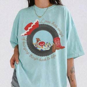 May include: A light blue t-shirt featuring a graphic of a black tire framing cartoon characters. The text reads Love has ways of bringing things back to life. A red cowboy hat and a red cowboy boot are positioned on the sides of the tire graphic.