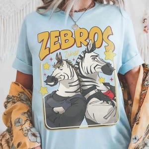 Op de afbeelding: Lichtblauw T-shirt met een cartoon illustratie van twee zebra's in pakken, met het woord "ZEBROS" in gele blokletters. De zebra's staan tegen een achtergrond van sterren en een gele rand. Het woord "Zebras" is cursief geschreven.