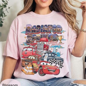 May include: A pale pink t-shirt with a graphic of Cars characters, including Lightning McQueen, and the words "Radiator Springs". The design includes a trophy and chequered flags. The shirt has a loose fit.