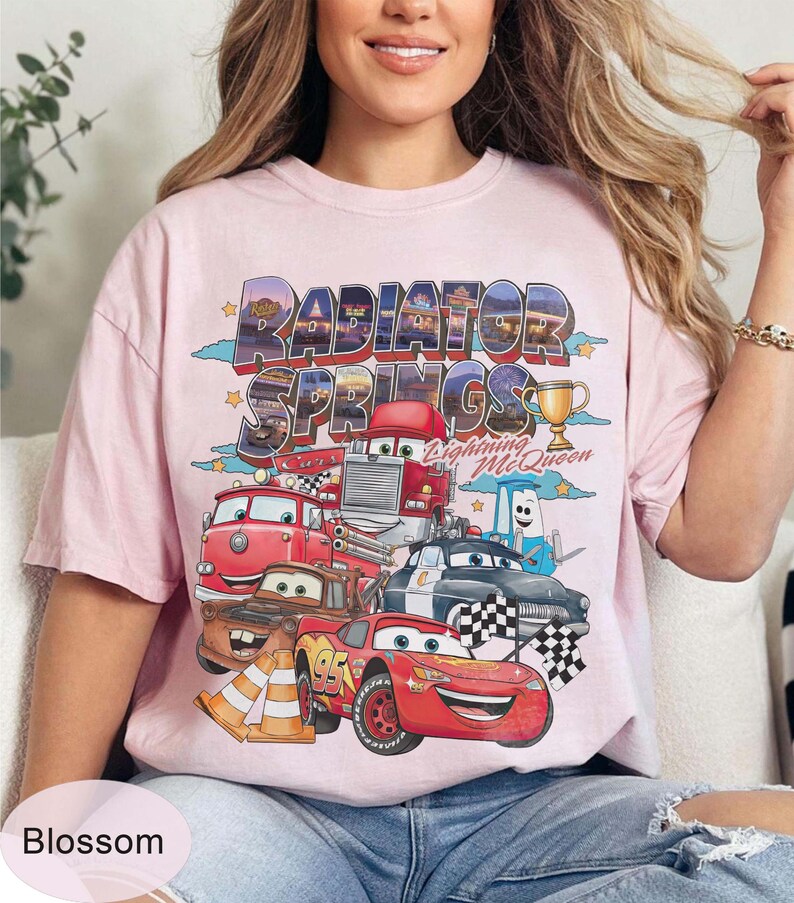 May include: A pale pink t-shirt with a graphic of Cars characters, including Lightning McQueen, and the words "Radiator Springs". The design includes a trophy and chequered flags. The shirt has a loose fit.