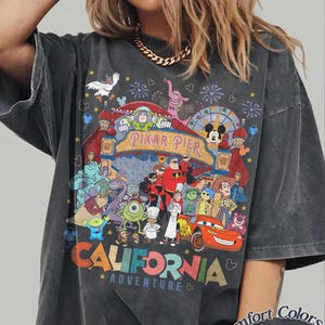 May include: Dark gray t-shirt featuring a colorful graphic of Pixar characters and the text "Pixar Pier" and "California Adventure". The shirt is labeled "Comfort Colors Pepper".