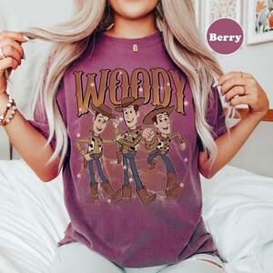 Op de afbeelding: Bessenkleurig T-shirt met het woord "WOODY" in een gouden, western-stijl lettertype boven een afbeelding van drie Woody-stripfiguren. De figuren dragen cowboyhoeden, vesten en laarzen. Het shirt heeft een vintage, distressed look.
