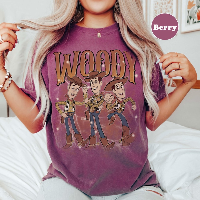 Op de afbeelding: Bessenkleurig T-shirt met het woord "WOODY" in een gouden, western-stijl lettertype boven een afbeelding van drie Woody-stripfiguren. De figuren dragen cowboyhoeden, vesten en laarzen. Het shirt heeft een vintage, distressed look.