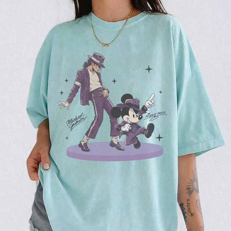 Pode incluir: Uma t-shirt azul-clara com uma estampa gr&aacute;fica de Michael Jackson e Mickey Mouse, ambos vestindo ternos e chap&eacute;us fedora roxos. O design inclui o texto Michael Jackson e Mickey Mouse com pequenos detalhes de estrelas numa plataforma circular.