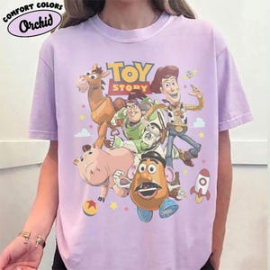 May include: A light purple t-shirt featuring a graphic print of Toy Story characters including Woody, Buzz Lightyear, Bullseye, Hamm, and Mr. Potato Head. The text "TOY STORY" is printed in yellow and red above the characters with small stars and a rocket.