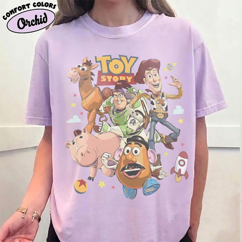 May include: A light purple t-shirt featuring a graphic print of Toy Story characters including Woody, Buzz Lightyear, Bullseye, Hamm, and Mr. Potato Head. The text "TOY STORY" is printed in yellow and red above the characters with small stars and a rocket.