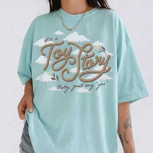 Pu&ograve; includere: Una t-shirt azzurro chiaro con una grafica di nuvole bianche e il testo "it's a Toy Story" scritto in un carattere marrone stile corda, con il testo "Baby, just say "yes"" stampato sotto in corsivo marrone scuro.