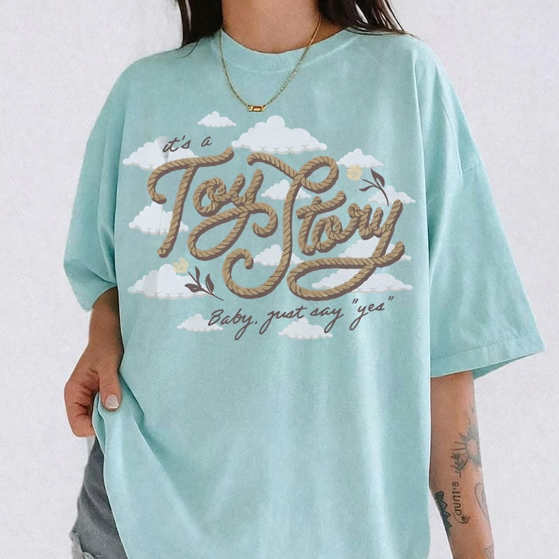 Pu&ograve; includere: Una t-shirt azzurro chiaro con una grafica di nuvole bianche e il testo "it's a Toy Story" scritto in un carattere marrone stile corda, con il testo "Baby, just say "yes"" stampato sotto in corsivo marrone scuro.