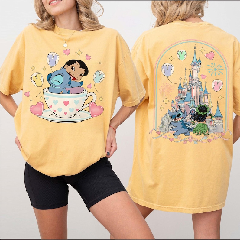 Puede incluir: Camiseta amarilla mostaza con un dise&ntilde;o de dibujos animados de Stitch y Lilo. El frente muestra a Stitch abrazando a Lilo en una taza de t&eacute; con corazones y globos. El dise&ntilde;o trasero presenta a Stitch y Lilo frente a un castillo con fuegos artificiales y globos.