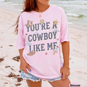May include: A light pink short-sleeve t-shirt featuring the dark blue text "YOU'RE A COWBOY LIKE ME" printed on the front. The design includes illustrations of cowboy boots, a hat, a star, a rocket, clouds, and a ball in various colors.