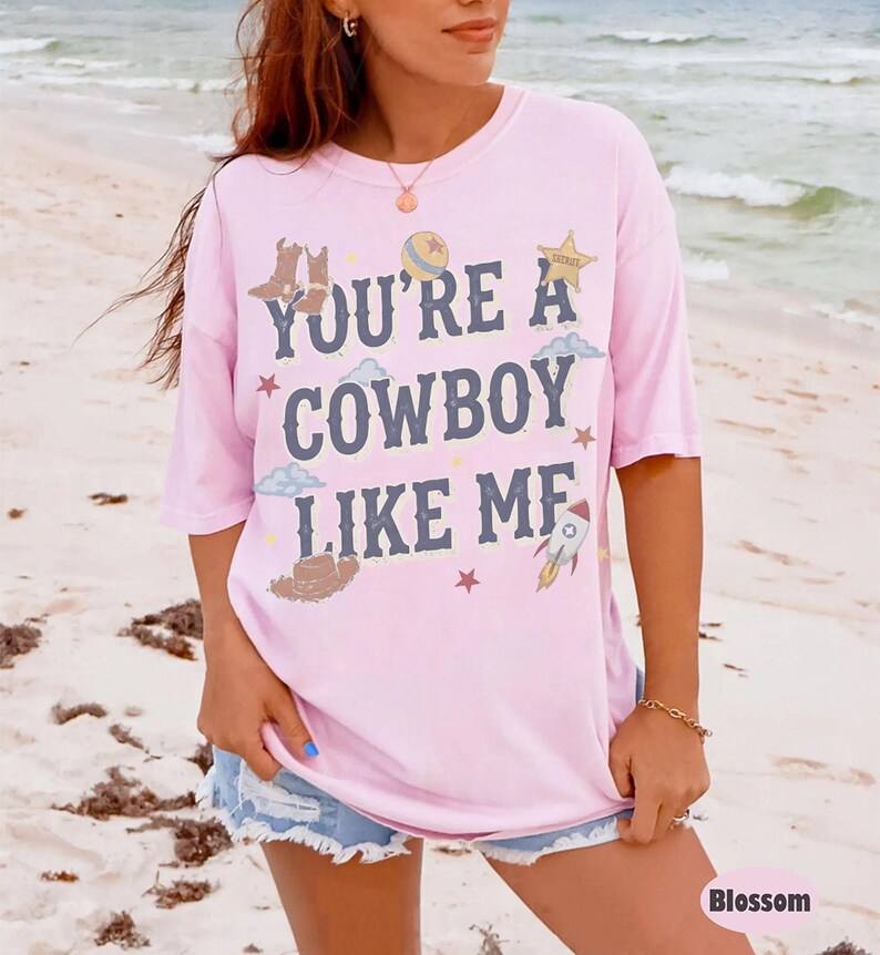 May include: A light pink short-sleeve t-shirt featuring the dark blue text "YOU'RE A COWBOY LIKE ME" printed on the front. The design includes illustrations of cowboy boots, a hat, a star, a rocket, clouds, and a ball in various colors.