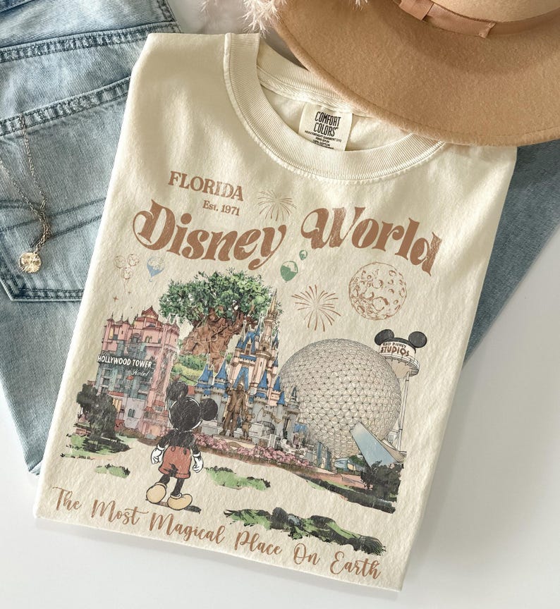 Może przedstawiać: Kremowa koszulka z grafiką Disney World na Florydzie. Projekt zawiera Myszka Miki, Hollywood Tower, zamek Kopciuszka i Spaceship Earth. Tekst na koszulce brzmi "Disney World" i "The Most Magical Place On Earth."