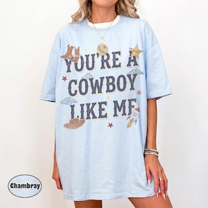 May include: A light blue t-shirt featuring the dark blue text "YOU'RE A COWBOY LIKE ME" printed on the front. The design includes graphic illustrations of cowboy boots, a hat, a star, a planet, and a rocket ship in muted earth tones.