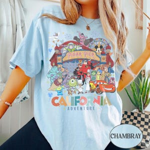 May include: Light blue t-shirt featuring a colorful graphic of Pixar Pier characters and the text "CALIFORNIA ADVENTURE". The design includes characters from various Pixar movies. The shirt has short sleeves and a relaxed fit.