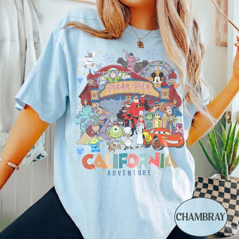 May include: Light blue t-shirt featuring a colorful graphic of Pixar Pier characters and the text "CALIFORNIA ADVENTURE". The design includes characters from various Pixar movies. The shirt has short sleeves and a relaxed fit.