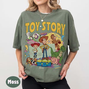 May include: Moss green t-shirt with the words "TOY STORY" in yellow and a star. The shirt features cartoon characters such as Buzz Lightyear, Woody, Jessie, and others. The t-shirt has a relaxed fit.