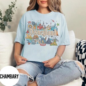 May include: Light blue t-shirt with a colorful Disneyland Resort graphic. The design features iconic park landmarks and the Disneyland logo. The shirt is a relaxed fit and has short sleeves. The overall style is casual and suitable for everyday wear.