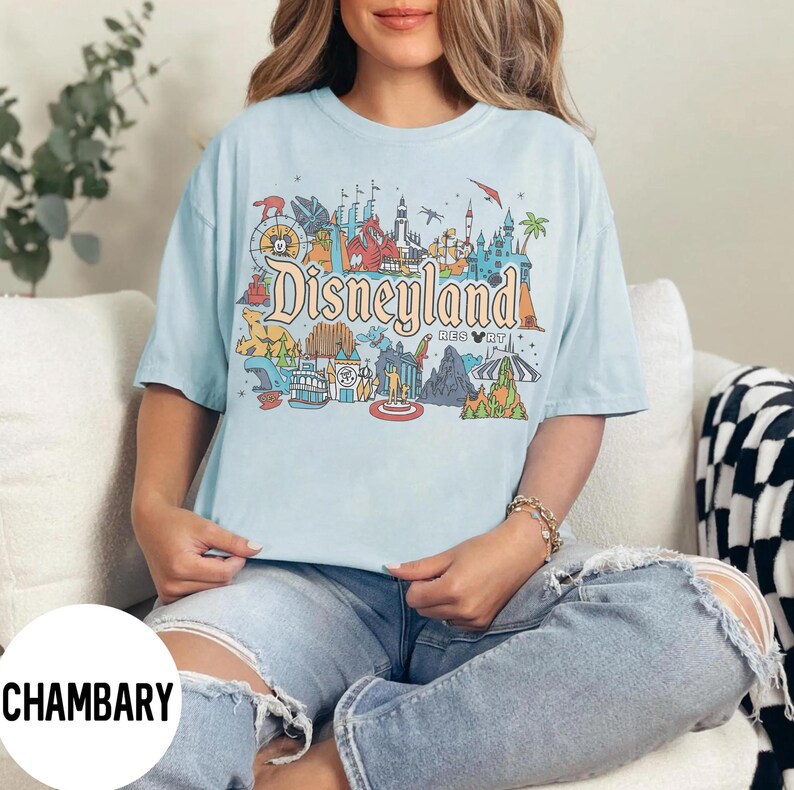 May include: Light blue t-shirt with a colorful Disneyland Resort graphic. The design features iconic park landmarks and the Disneyland logo. The shirt is a relaxed fit and has short sleeves. The overall style is casual and suitable for everyday wear.
