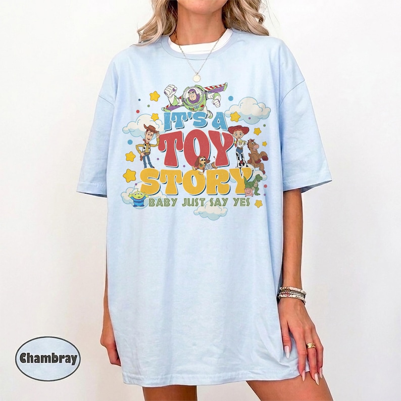 May include: A light blue t-shirt featuring a graphic print of Toy Story characters including Woody, Buzz Lightyear, Jessie, and Bullseye. The text reads IT'S A TOY STORY BABY JUST SAY YES, surrounded by yellow stars and white clouds on the front.