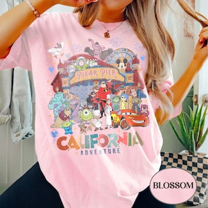 May include: A light pink t-shirt featuring a colorful graphic of various Disney and Pixar characters, including Buzz Lightyear, Mickey Mouse, and Lightning McQueen. The text "Pixar Pier" and "California Adventure" are also visible. The shirt has short sleeves.