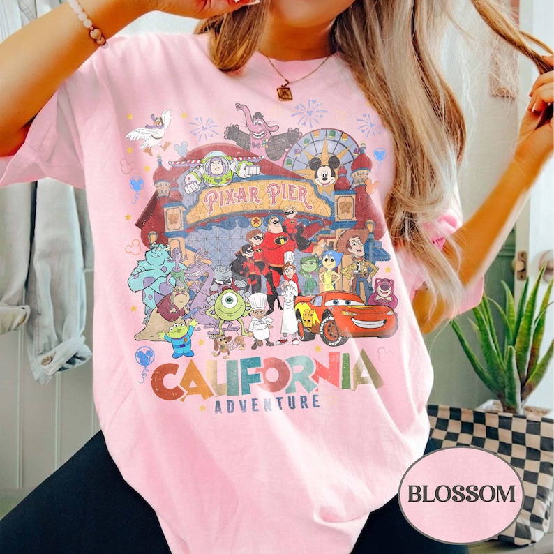 May include: A light pink t-shirt featuring a colorful graphic of various Disney and Pixar characters, including Buzz Lightyear, Mickey Mouse, and Lightning McQueen. The text "Pixar Pier" and "California Adventure" are also visible. The shirt has short sleeves.
