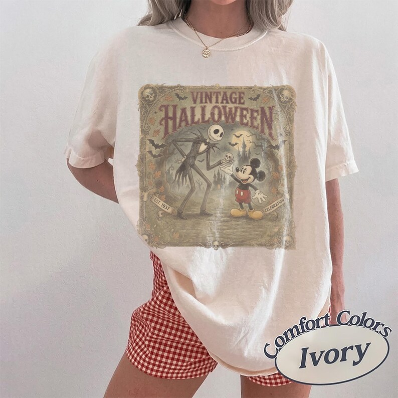 May include: An ivory short-sleeve t-shirt featuring a rectangular graphic with the text VINTAGE HALLOWEEN, EST. 1937, and CELEBRATION. The illustration depicts Jack Skellington and Mickey Mouse in a spooky landscape with bats and a castle.