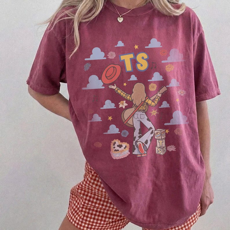 Op de afbeelding: Een bordeauxrood t-shirt met korte mouwen en een grafische print van een persoon die gitaar speelt, een hoed, wolken, sterren, bloemen, een hartvormige taart en blokken met de tekst TS. De persoon draagt een geruit overhemd en een broek.