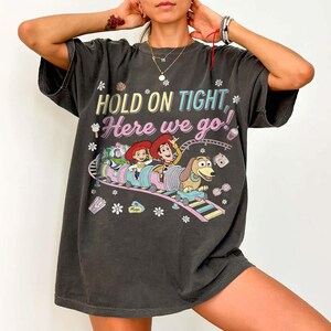 May include: Dark gray t-shirt featuring a colorful graphic of characters on a roller coaster with the text "HOLD ON TIGHT, Here we go!". The design includes Woody, Jessie, Buzz Lightyear, and Slinky Dog. The shirt has a vintage, worn look.