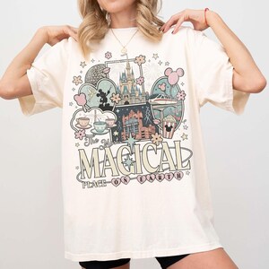 Puede incluir: Camiseta de color crema con un gr&aacute;fico colorido con el texto "The Most Magical Place on Earth". El dise&ntilde;o incluye ilustraciones de un castillo, tazas de t&eacute; y globos de Mickey Mouse. La camiseta tiene mangas cortas.