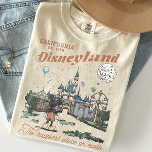Puede incluir: Camiseta color crema con un gr&aacute;fico de Disneyland. El dise&ntilde;o presenta un castillo, Mickey Mouse, una noria y el texto "California Disneyland Est. 1955" y "The happiest place on earth."
