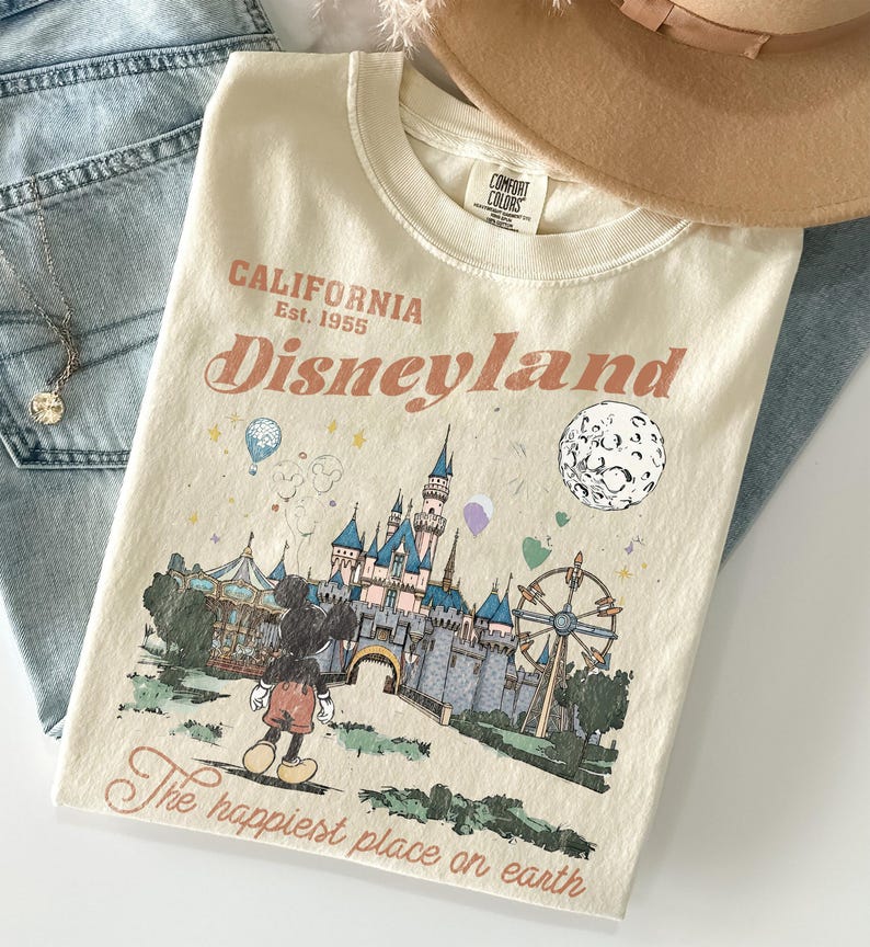 Puede incluir: Camiseta color crema con un gr&aacute;fico de Disneyland. El dise&ntilde;o presenta un castillo, Mickey Mouse, una noria y el texto "California Disneyland Est. 1955" y "The happiest place on earth."