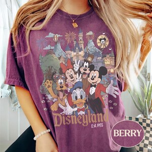 Puede incluir: Camiseta de color bayas con un gr&aacute;fico de Disneyland con Mickey Mouse, Minnie Mouse, Donald Duck y Goofy. El gr&aacute;fico incluye un castillo, fuegos artificiales y el texto "Disneyland Est. 1955". La camiseta tiene mangas cortas y cuello redondo.