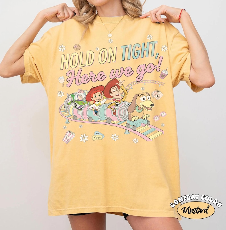 May include: Mustard-colored t-shirt with the text "HOLD ON TIGHT, Here we go!" and a graphic of Toy Story characters on a roller coaster. The shirt also has the words "Comfort Color" and "Mustard" in a circle.