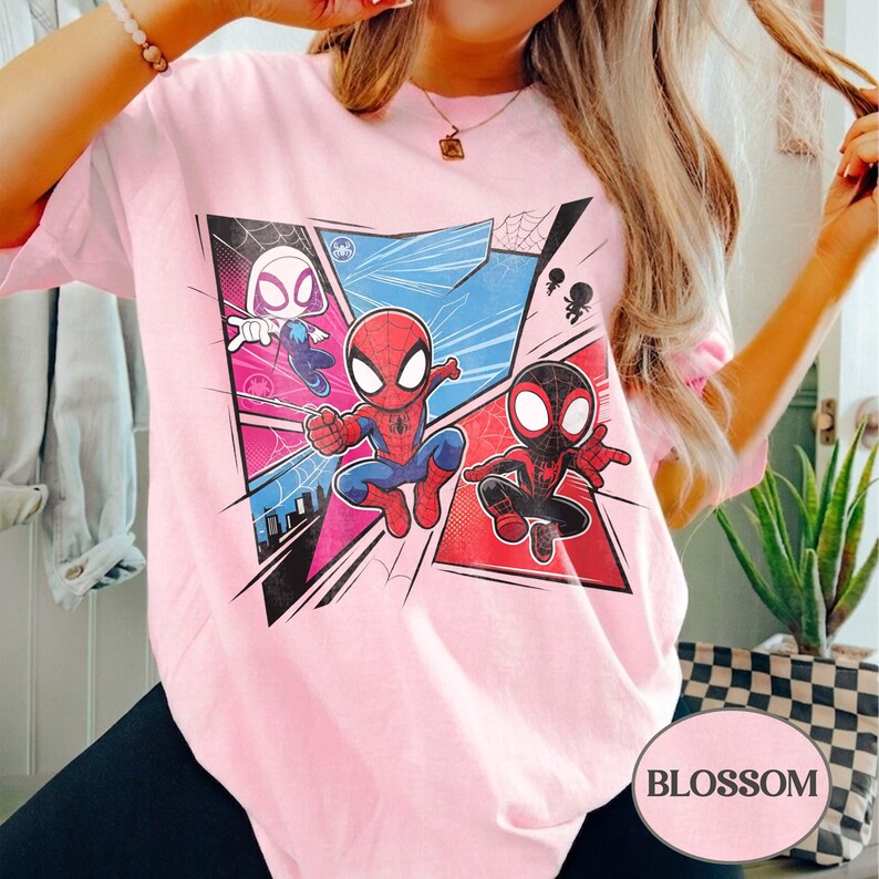 K&ouml;nnte beinhalten: Hellrosa T-Shirt mit einem farbenfrohen Grafikdesign von Spider-Man, Spider-Gwen und Miles Morales. Das Design beinhaltet Comic-Panels mit blauen, roten und rosa Hintergr&uuml;nden. Das Wort "BLOSSOM" ist unten rechts aufgedruckt.