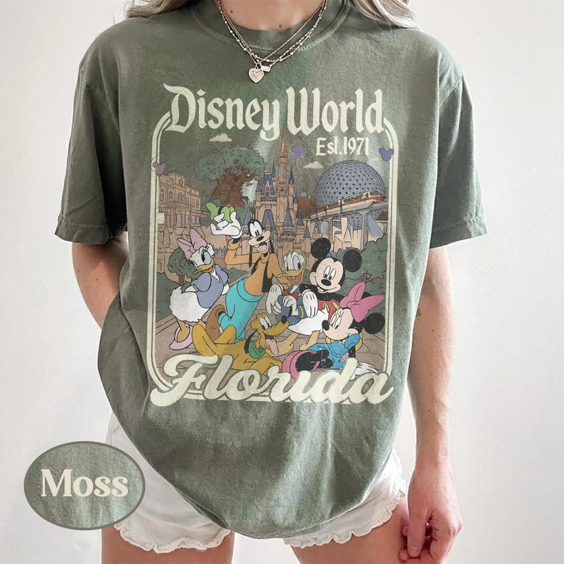 May include: Moss green t-shirt featuring a Disney World graphic. The design includes Mickey and Minnie Mouse, Donald and Daisy Duck, Goofy, and Pluto, with the text "Disney World Florida Est. 1971". The shirt has a crew neck and short sleeves.