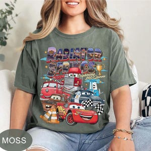 May include: Moss green t-shirt with a vibrant graphic of Cars movie characters, including Lightning McQueen and the text "Radiator Springs." The design incorporates a trophy and chequered flags.