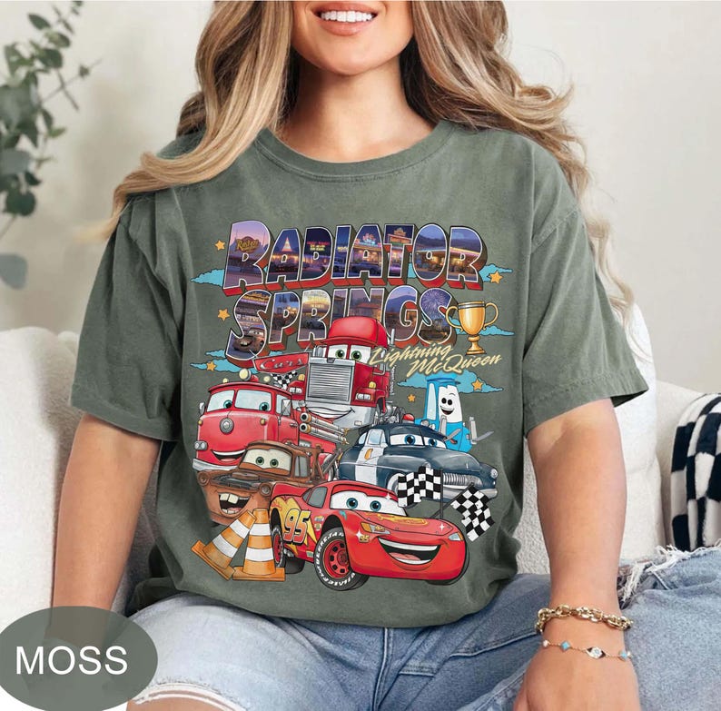 May include: Moss green t-shirt with a vibrant graphic of Cars movie characters, including Lightning McQueen and the text "Radiator Springs." The design incorporates a trophy and chequered flags.