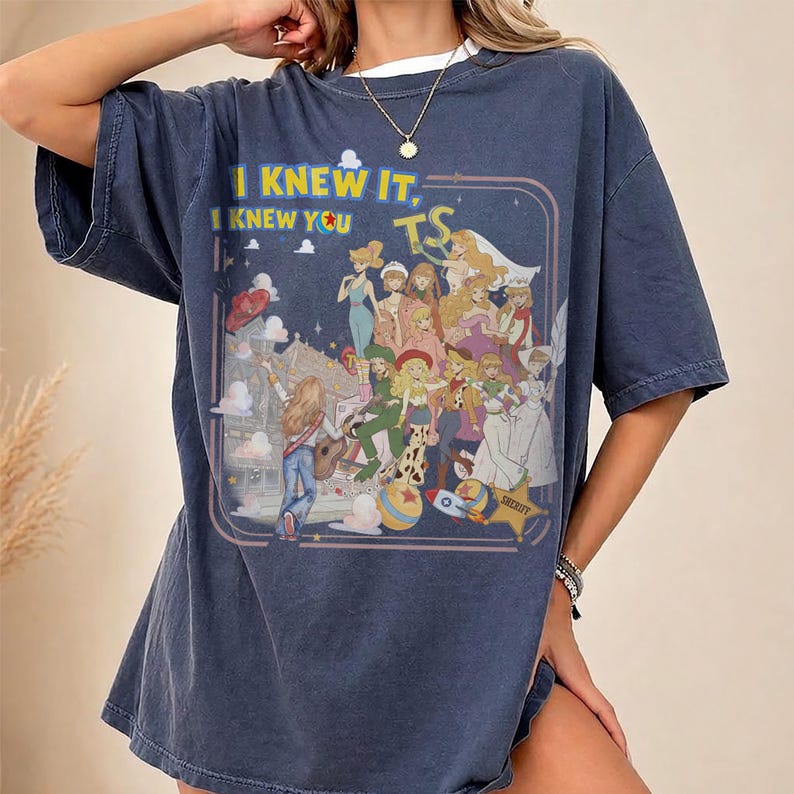 May include: A dark blue short-sleeve t-shirt featuring a graphic print of various animated characters. The text "I KNEW IT, I KNEW YOU" and "TS" is printed above the characters. The design includes a star, a rocket, and a ball in a retro style.