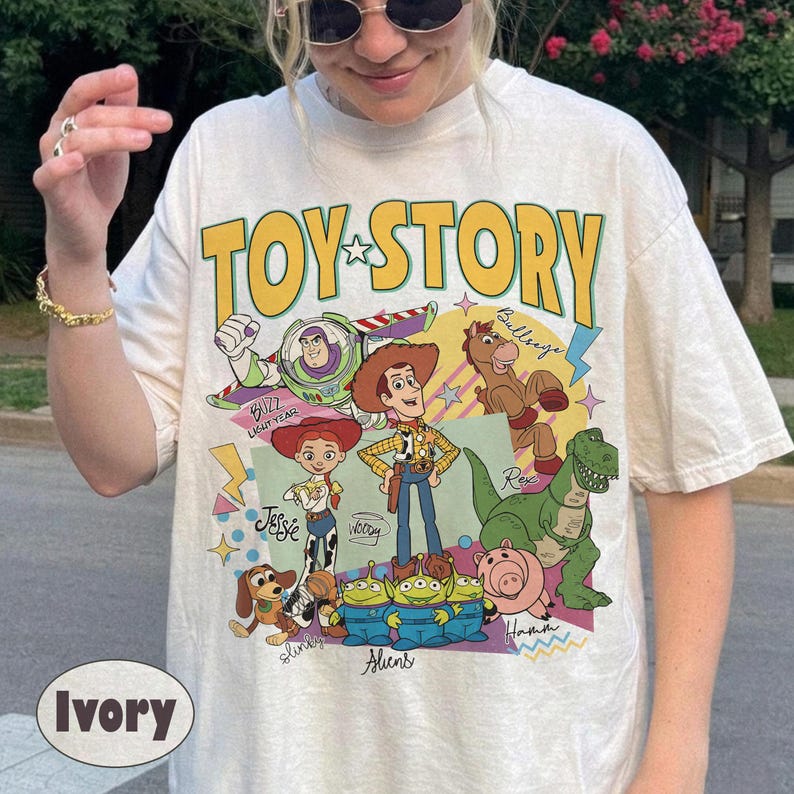 May include: Ivory-coloured t-shirt with the words "TOY STORY" in yellow, featuring cartoon characters including Buzz Lightyear and Woody. The shirt has a crew neck and short sleeves.