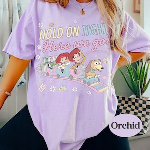 May include: A light purple t-shirt featuring a colorful graphic of characters from the movie Toy Story on a roller coaster. The text "HOLD ON TIGHT, Here we go!" is printed above the graphic. The shirt has short sleeves.