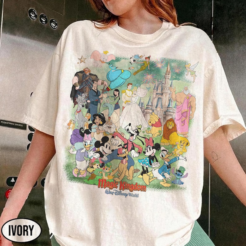 Może przedstawiać: Kremowa koszulka z kolorową grafiką postaci Disneya, w tym Myszki Miki, Myszki Minnie, Kaczora Donalda i innych. Grafika zawiera tekst "Magic Kingdom Our Disney World".
