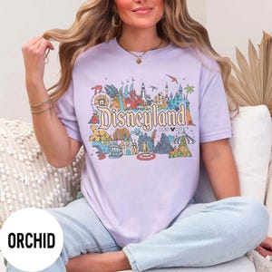 May include: Orchid-colored t-shirt featuring a colorful Disneyland graphic. The design includes iconic park elements and the Disneyland logo. The shirt is a relaxed fit and has short sleeves.