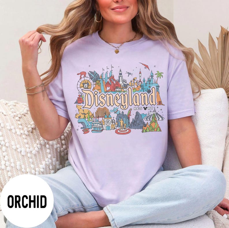 May include: Orchid-colored t-shirt featuring a colorful Disneyland graphic. The design includes iconic park elements and the Disneyland logo. The shirt is a relaxed fit and has short sleeves.