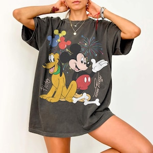 May include: Dark gray t-shirt featuring Mickey Mouse, Pluto, and fireworks. Mickey Mouse is wearing red shorts and white gloves. Pluto is yellow with a green collar. The shirt has the words "Mickey Mouse" and "Pluto" printed on it.