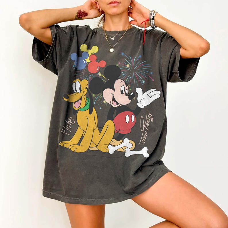 May include: Dark gray t-shirt featuring Mickey Mouse, Pluto, and fireworks. Mickey Mouse is wearing red shorts and white gloves. Pluto is yellow with a green collar. The shirt has the words "Mickey Mouse" and "Pluto" printed on it.