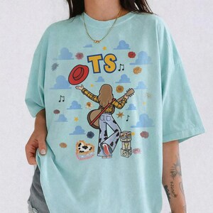 Op de afbeelding: Een lichtblauw t-shirt met een afbeelding van een persoon met lang bruin haar die gitaar speelt, gekleed in een geel geruit overhemd en een broek met koeienprint. Het ontwerp bevat de tekst TS, een rode hoed en blauwe wolken.