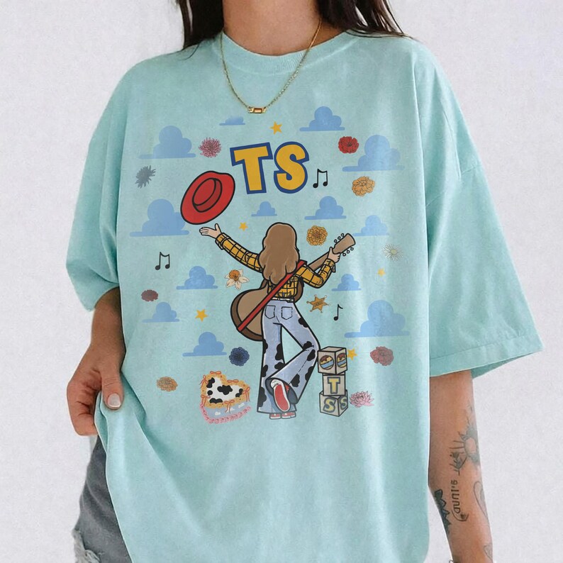 Op de afbeelding: Een lichtblauw t-shirt met een afbeelding van een persoon met lang bruin haar die gitaar speelt, gekleed in een geel geruit overhemd en een broek met koeienprint. Het ontwerp bevat de tekst TS, een rode hoed en blauwe wolken.