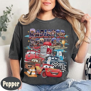 May include: Dark grey t-shirt with a colourful graphic of Cars characters, including Lightning McQueen. The text "Radiator Springs" is above the cars. A gold trophy is also shown on the shirt.
