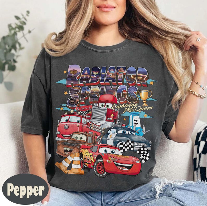 May include: Dark grey t-shirt with a colourful graphic of Cars characters, including Lightning McQueen. The text "Radiator Springs" is above the cars. A gold trophy is also shown on the shirt.